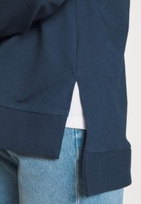 Felpa blu navy con orlo a coste, fenditure laterali e tessuto testurizzato, indossata sopra una camicia bianca, abbinata a jeans in denim blu chiaro.