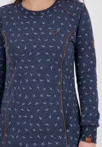 Sudadera de color azul marino con estampado de pájaros en azul claro, con costuras marrones, un logo de corazón y puños acanalados. Tela suave y texturada.