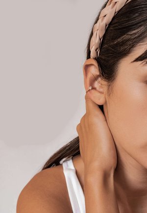 Geflochtener beiger Stoffhaarband, der auf dunklem Haar getragen wird. Nahaufnahme einer kleinen goldenen Creole mit floralem Detail am Ohr. Glatte Hautstruktur.