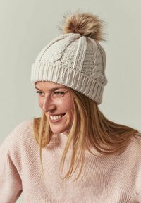 Gebreide crèmekleurige beanie met een dikke geribbelde band en een gestructureerd patroon; heeft een pluizige pompon bovenop.
