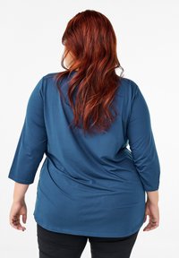 Blue, long-sleeve top with a relaxed fit and rounded hem. Smooth texture, subtle stitching details, worn with black pants, hair flowing over back.