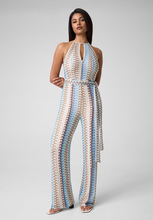 Femme debout, les bras croisés dans le dos, portant une combinaison sans manches col halter avec des motifs verticaux bleu pastel, beige et blanc.