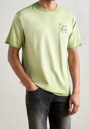 Man draagt een lichtgroen T-shirt met korte mouwen met de tekst "Club Soda Scotch & Soda" op de borst en donkergrijze jeans, hand in de zak.