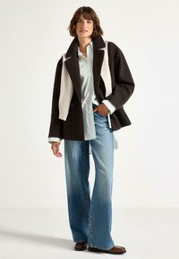 Cappotto oversize in misto lana marrone e crema, abbinato a una camicia a righe blu chiaro e jeans a gamba larga sbiaditi. Accentuato da stivali marroni.