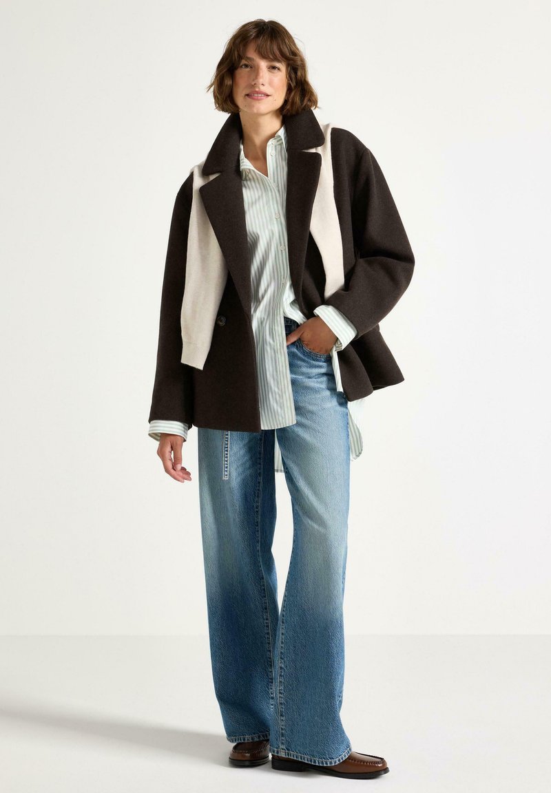 Cappotto oversize in misto lana marrone e crema, abbinato a una camicia a righe blu chiaro e jeans a gamba larga sbiaditi. Accentuato da stivali marroni.