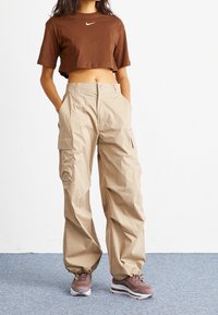 Camiseta corta marrón con un logo blanco de Nike, combinada con pantalones cargo beiges con bolsillos laterales y un corte relajado, llevados con zapatillas marrones.