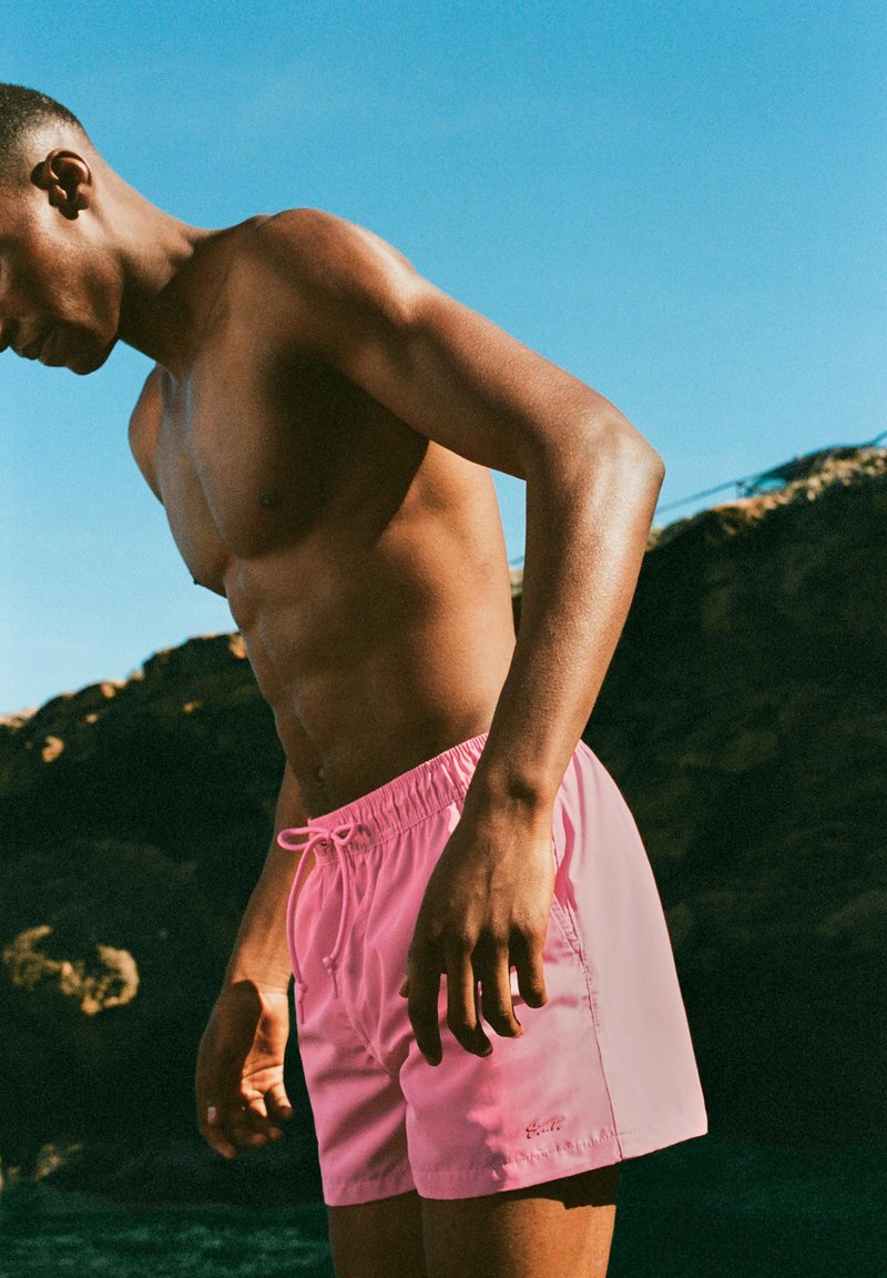 Homme torse nu portant un short de bain rose se tient près d’une côte rocheuse sous un ciel bleu clair.