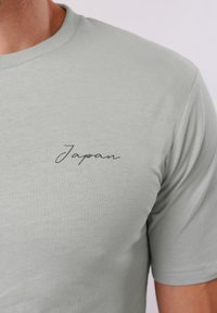 Gros plan d'une personne portant un t-shirt gris clair avec le mot « Japan » imprimé en petites cursives noires sur le côté gauche de la poitrine.