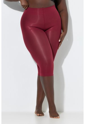 Kurvige Frau steht barfuß an einer weißen Wand und trägt hoch taillierte, glänzende kniehohe Leggings in Burgunderrot.