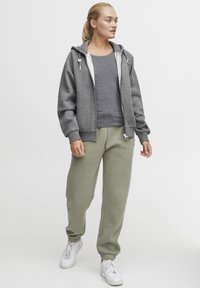 Gray zip-up hoodie with drawstring, paired with a gray top and olive green sweatpants. Soft fabric, casual fit, and white sneakers.