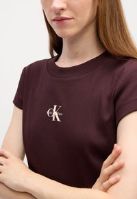 Bordowa koszulka z prążkowanej tkaniny, z okrągłym dekoltem i krótkimi rękawami, z kremowym logo Calvin Klein na środku klatki piersiowej.