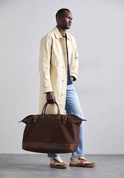 Brown duffle bag with leather accents and a textured bottom, carried by a man in a light beige coat, blue jeans, and colorful sneakers.