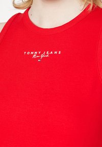 Haut rouge sans manches avec inscription "TOMMY JEANS New York" et petit logo Tommy Hilfiger sur le devant, porté par une personne à la peau claire et aux cheveux blonds.