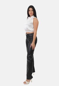 Donna con lunghi capelli neri che indossa un top in satin bianco senza maniche, pantaloni in pelle nera e sandali bianchi con tacco alto, in piedi di lato.