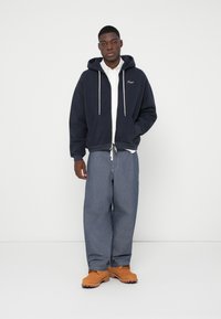 Μπλε σκούρο φούτερ με κουκούλα και κορδόνια, με μπροστινές τσέπες, φορεμένο πάνω από λευκό πουκάμισο με κουμπιά, συνδυασμένο με φαρδιά μπλε τζιν και ταμπά μπότες.