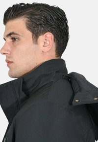 Giacca nera con colletto alto, caratterizzata da una combinazione di materiali, texture liscia e dettagli hardware visibili. Vista laterale.