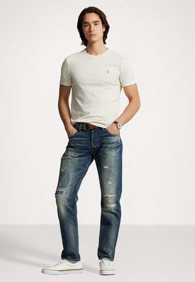 Hvit t-skjorte med kort erm og lite logo, slitte blå jeans med lapper, og hvite joggesko. Avslappet antrekk med minimale tilbehør.