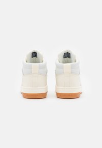 Wysokie trampki z kremową cholewką, szarymi akcentami i brązową gumową podeszwą. Z tyłu znajduje się logo oraz pętelka ułatwiająca zakładanie.