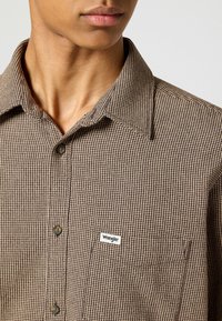 Brązowa koszula w kratę na guziki z kołnierzykiem, z kieszenią na lewej piersi i małą białą metką z logo Wrangler.