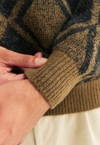Una mano che regola il polsino di un maglione lavorato a maglia con un motivo marrone e blu scuro, con dei pantaloni beige visibili sotto.