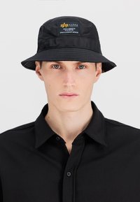 Black bucket hat made from a smooth fabric, featuring a logo patch on the front. Worn with a black button-up shirt.