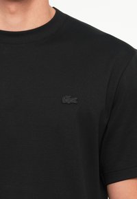 T-shirt en coton noir avec un col rond et des manches courtes. Présente un petit logo brodé noir sur la poitrine gauche. Texture douce.