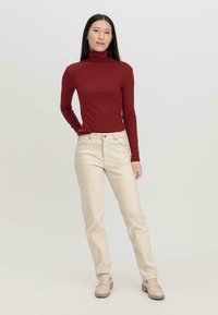 Langarm-Burgunder-Turtleneck-Oberteil, kombiniert mit cremefarbenen gerade geschnittenen Hosen und hellbraunen Loafers auf einem neutralen Hintergrund.