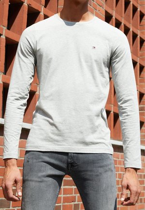 Light gray long-sleeve t-shirt, cotton material, features a small logo on the chest; paired with dark gray slim-fit jeans. Textured brick background.