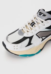 Zapatilla blanca con parte superior de malla, acentos en gris y negro, punta redonda y suela gruesa en crema y verde azulado. Presenta cordones y detalles perforados.