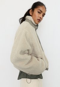 Kraftig beige fleecejakke med grå krave, oversize pasform og sidelommer. Båret over en sort-hvid ternet skjorte.