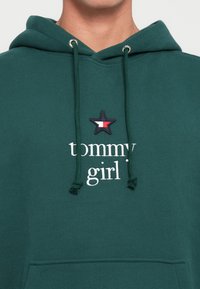 Felpa verde in cotone con tasca frontale. Caratterizzata da un logo bianco con il testo "tommy girl" e un toppa a forma di stella nera con un design rosso e bianco.