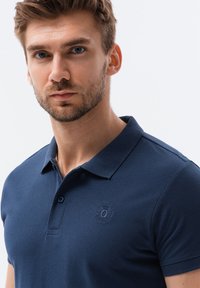 Marinblå pikétröja i strukturerad tyg, med krage, två knappar och en broderad logotyp på bröstet.