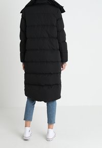 Abrigo acolchado negro con un diseño largo y voluminoso, con costuras horizontales, capucha y un dobladillo asimétrico, combinado con jeans azules y zapatillas blancas.