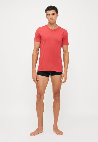 T-shirt rouge à manches courtes avec un col ras du cou, fabriqué à partir d'un matériau lisse, porté avec un short de boxe noir, mannequin pieds nus contre un fond blanc.