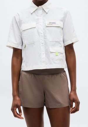 Personne portant une chemise blanche à manches courtes avec poches et un short marron sur un fond uni.