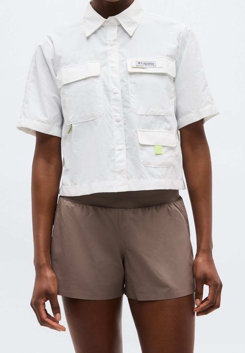 Personne portant une chemise blanche à manches courtes avec poches et un short marron sur un fond uni.