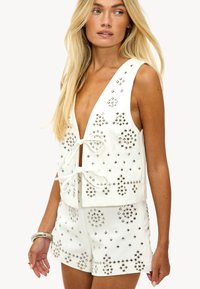 Mujer rubia que lleva un conjunto de chaleco sin mangas blanco y pantalones cortos, con adornos de tachuelas cuadradas plateadas y dos cierres delanteros con lazo.