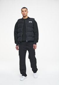 Black puffer vest with a zip front and logo, worn over a black sweatshirt. Paired with black cargo pants and white sneakers.