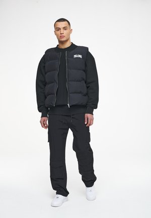 Black puffer vest with a zip front and logo, worn over a black sweatshirt. Paired with black cargo pants and white sneakers.