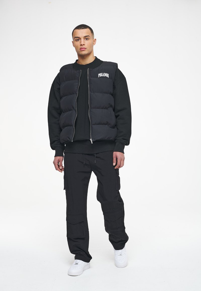 Black puffer vest with a zip front and logo, worn over a black sweatshirt. Paired with black cargo pants and white sneakers.