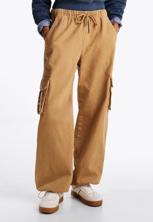 Khaki cargo bukser laget av myk fabric, med elastisk midje, to sidelommer og dekorativ lapp på venstre ben.