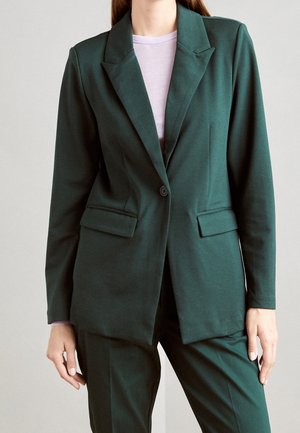 Mujer vestida con un blazer entallado verde oscuro y pantalones a juego, con una blusa violeta clara debajo, de pie contra un fondo plano.