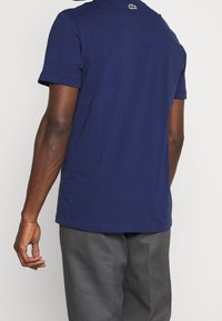 Navy blue cotton t-shirt with short sleeves, round neckline, and a small logo on the back, worn with gray pants. Smooth texture.