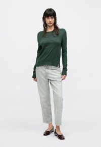 Γυναίκα που φοράει σκούρο πράσινο πουλόβερ με μακριά μανίκια, ανοιχτό γκρι τζιν και σκούρα κόκκινα μπαλαρίνα παπούτσια, στέκεται μπροστά σε λευκό φόντο.