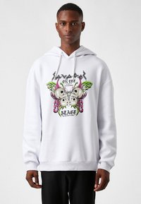 Sudadera blanca con un diseño gráfico que incluye calaveras, una mariposa y el texto "SAVAGE ON THE STAGE". Hecha de una tela suave con puños acanalados.