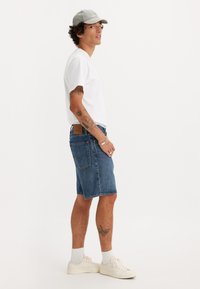 Jeansshorts in Mittelblau, knielang, mit einer Gesäßtasche, kombiniert mit einem schlichten weißen T-Shirt und einer grauen Kappe. Weiße Sneakers und kurze Socken.