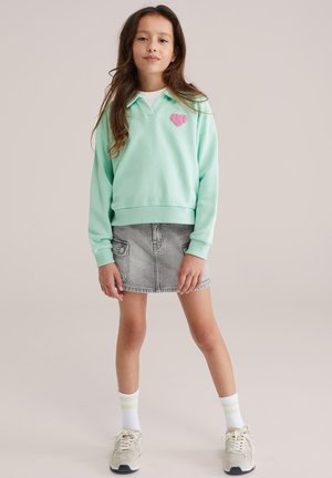 Mentolasto zeleni pulover s zakrpom u obliku srca, uparena s sivom traper suknjom. Bijele čarape s limetasto zelenim prugama i bež tenisice upotpunjuju odjevnu kombinaciju.