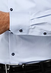 Lichtblauw overhemd met zwarte knopen, met een frisse textuur. Manchetdetails met knoopaccenten en een gladde afwerking. Zwarte leren riem zichtbaar.