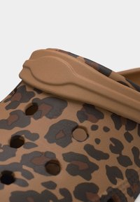 Gros plan sur une sabots à imprimé léopard marron et noir avec des trous de ventilation et une sangle arrière réglable de couleur beige.