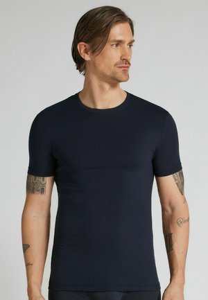 Man met middellang haar die een strak zwart T-shirt met korte mouwen draagt, kijkt naar rechts en toont tatoeages op beide armen.